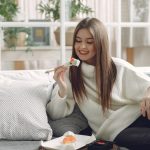 Smiling young lady in warm knitted sweater enjoying fresh sushi on wicker tray while relaxing on cozy sofa with soft cushions in modern living room